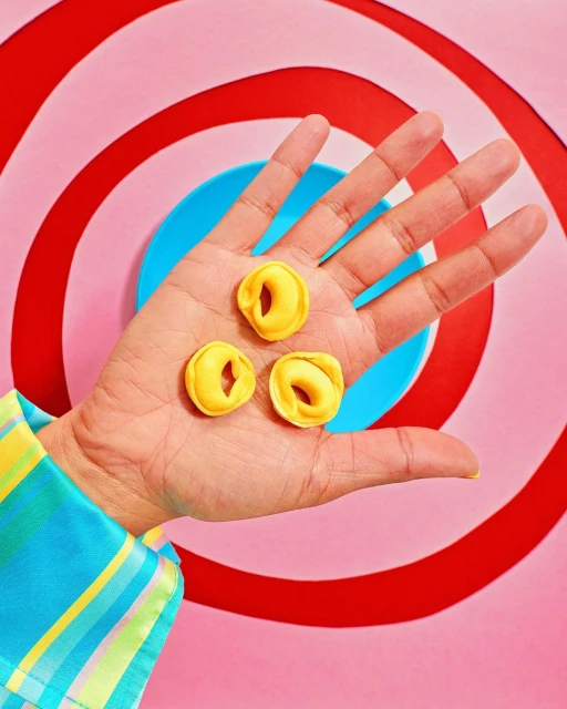 Three ring-shaped pasta pieces are held in an adult's hand against a colorful, graphic background. The sleeve is patterned with squares.