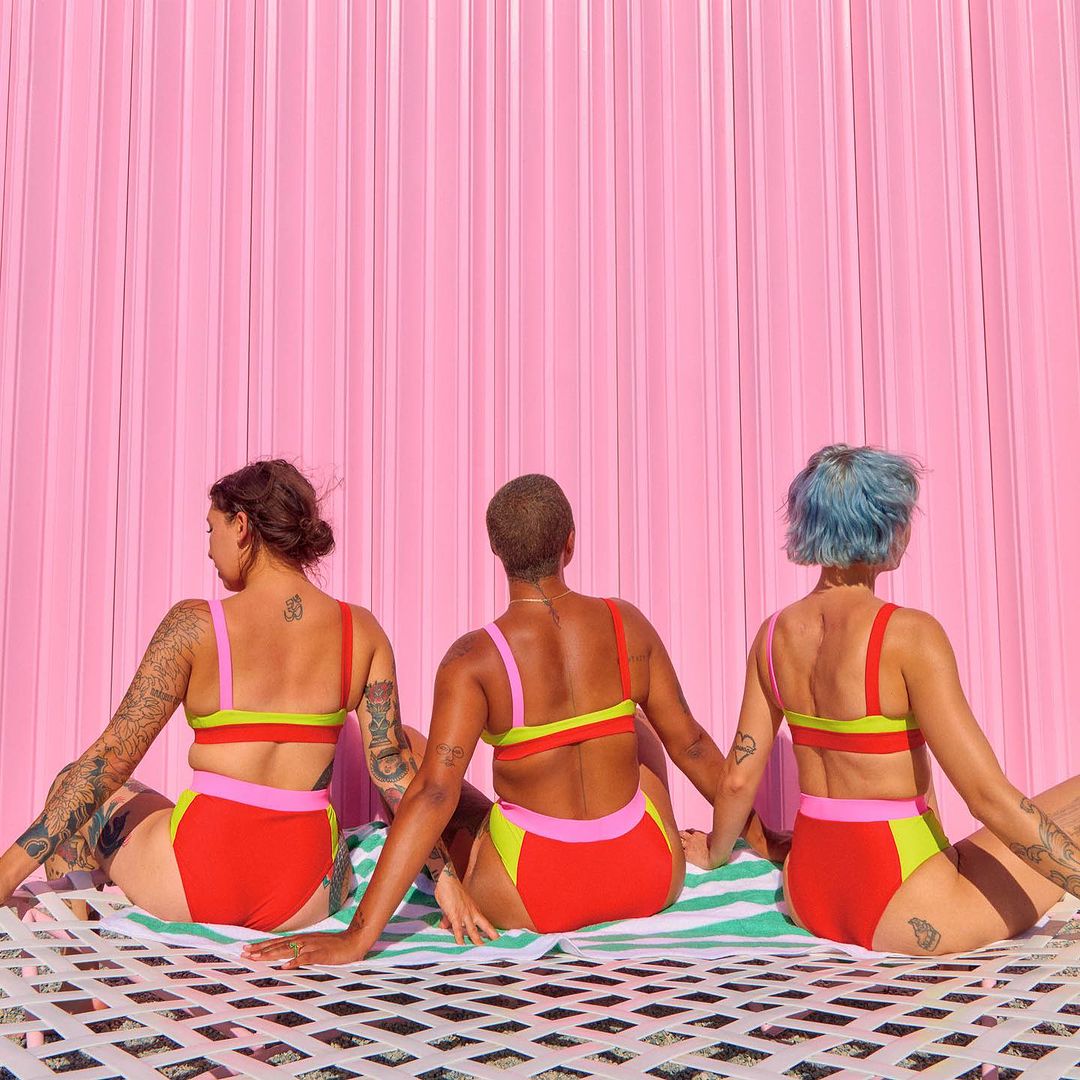 Three women in swimsuits sit on a white net over a striped towel against a pink background. The women face away from the camera.