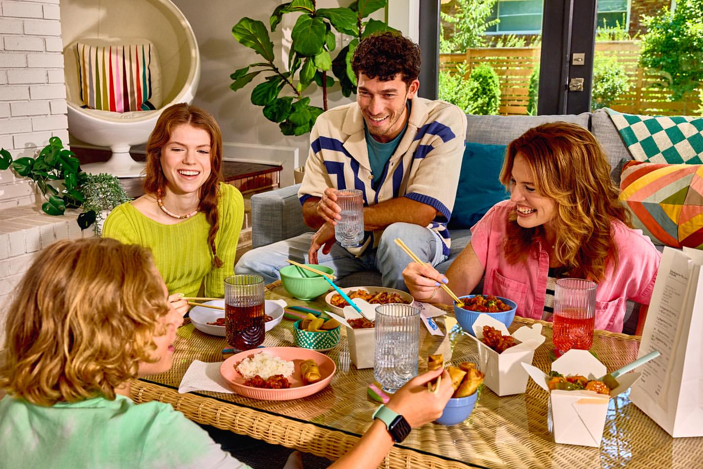 Four friends enjoy a casual meal at home, sitting around a wicker table with Chinese takeout, laughing and eating with chopsticks.