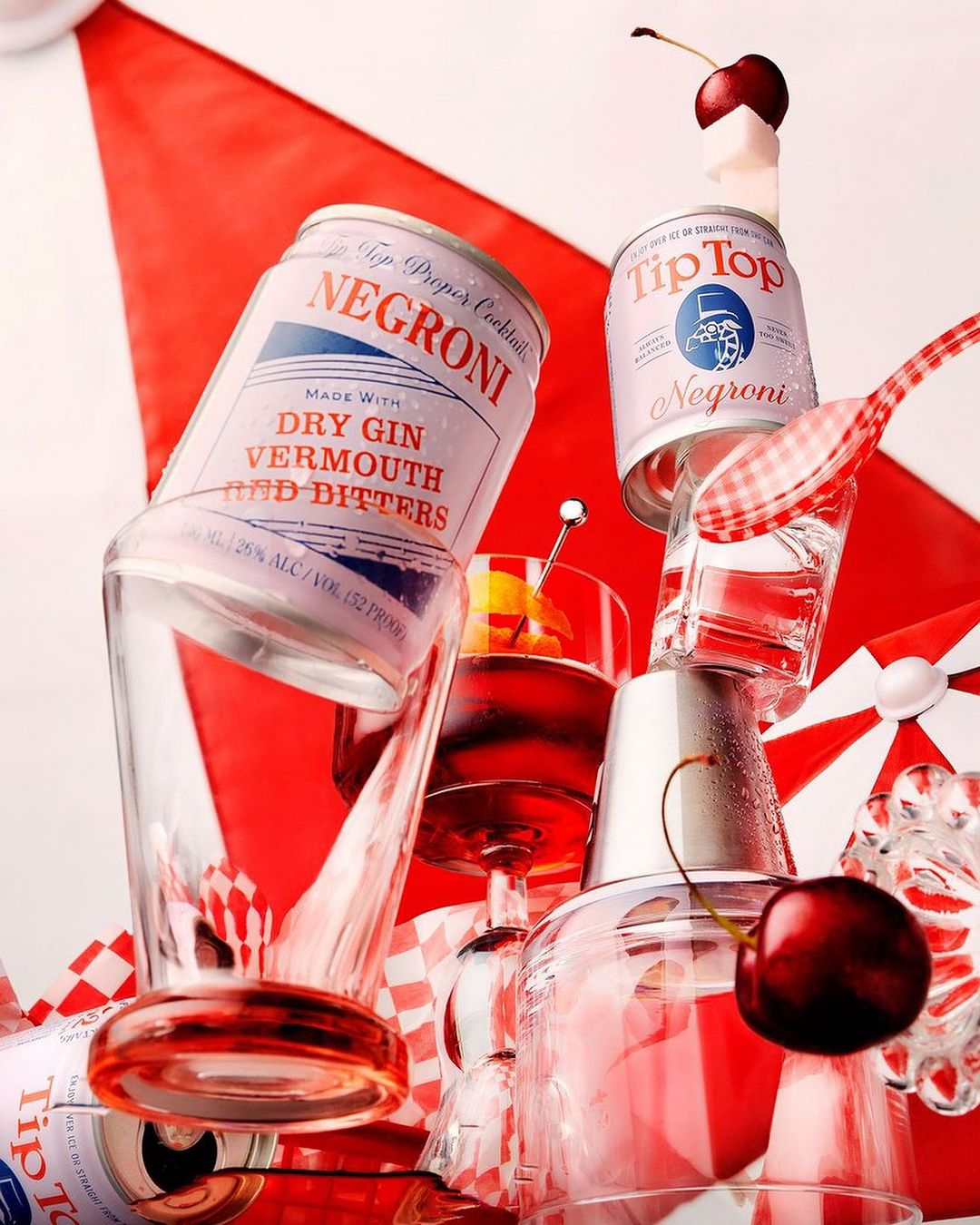Still life of two cans of Negroni cocktails stacked with cocktail glasses, garnish picks, and cherries against a red and white striped background.