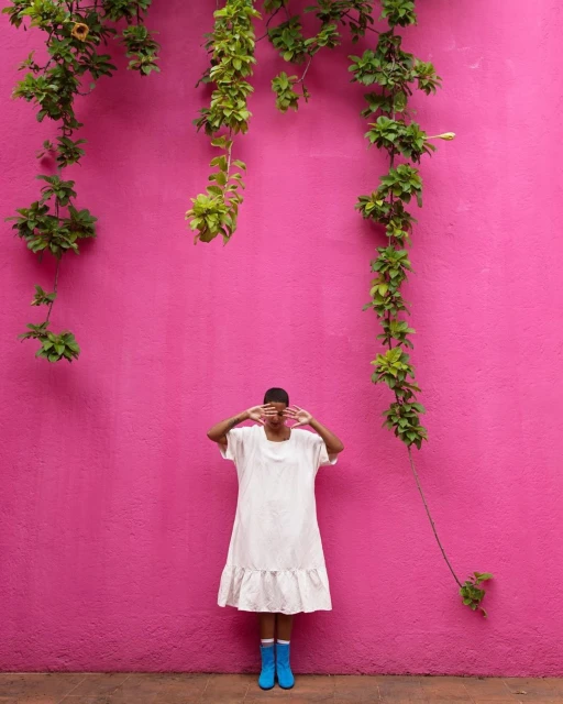 An adult woman with short hair poses with hands covering her face in front of a pink wall with green vines, wearing a white dress and blue boots.