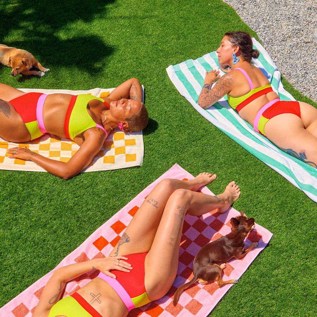 Three women in swimsuits sunbathing on patterned towels outdoors on green grass with two small dogs, enjoying a sunny day.