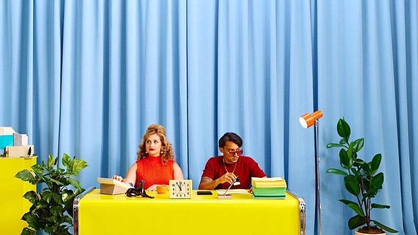 Two people sit at a yellow desk with vintage office objects, including a telephone, clock, and lamp, against a light blue curtain backdrop.