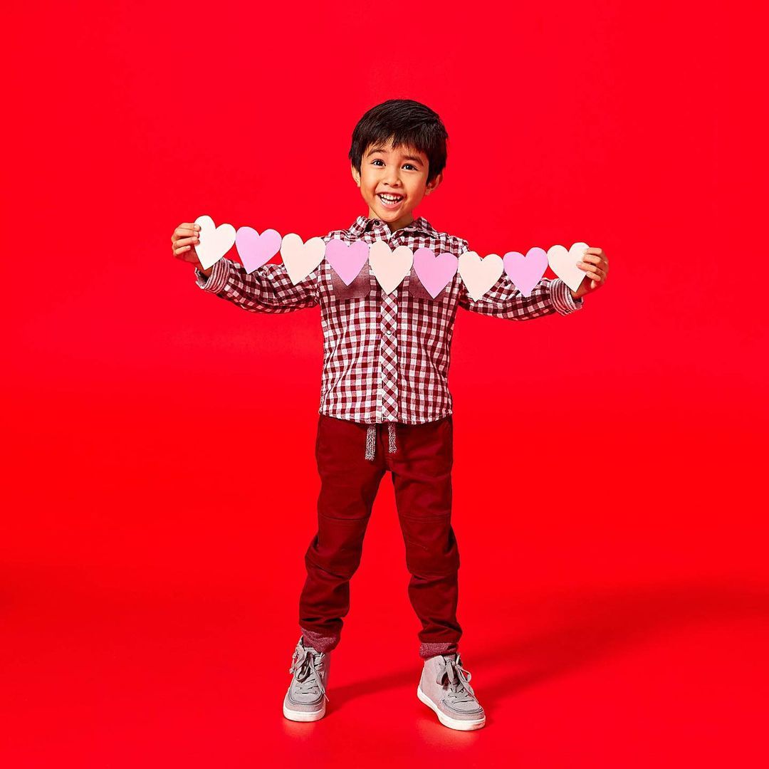 A young boy with dark hair smiles while holding a string of pastel paper hearts against a solid red studio backdrop, full shot.