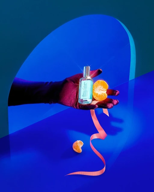 Product shot of a blue cologne bottle held by a gloved hand, presented with a mandarin orange against a blue backdrop with a peach ribbon.