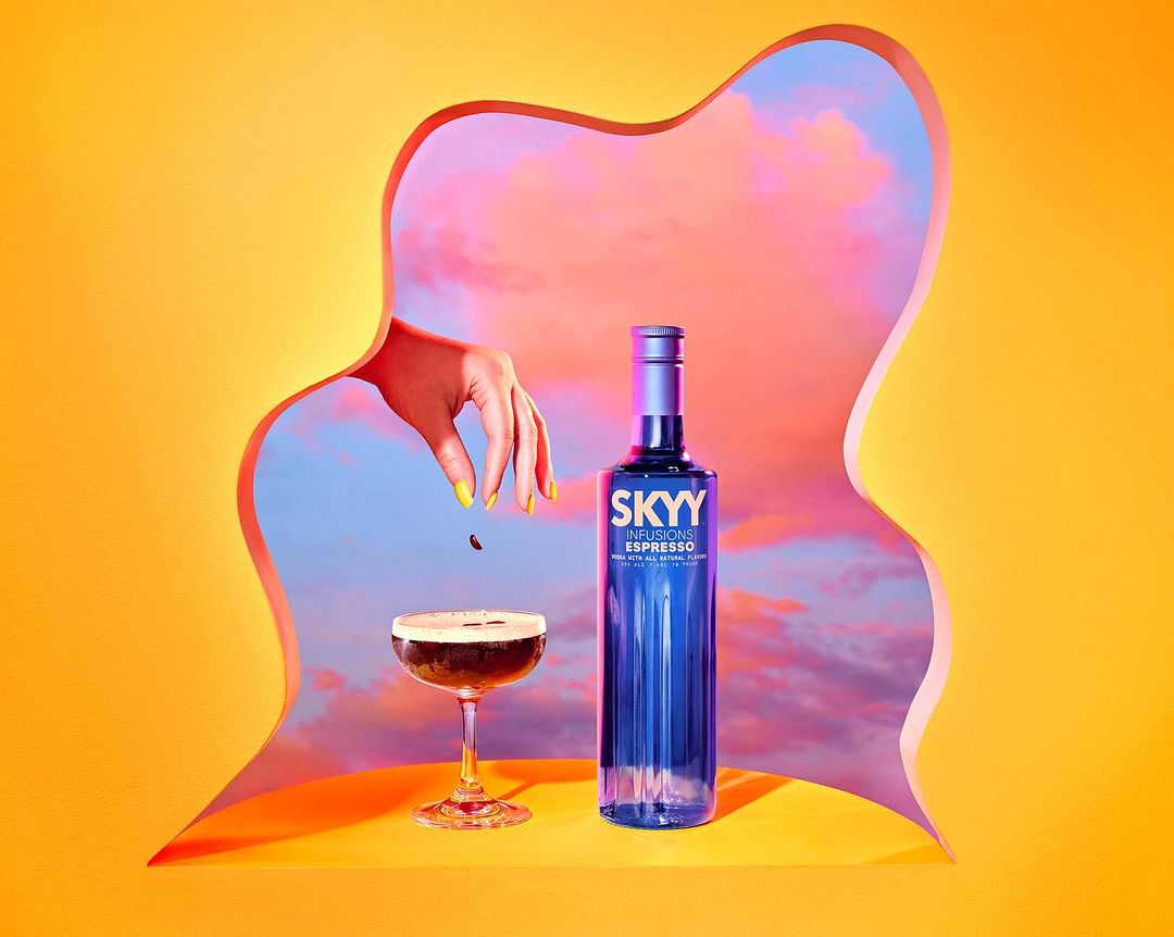 Vibrant studio shot of Skyy Infusions Espresso bottle, cocktail glass, and a hand dropping coffee bean against an orange background with cutout.