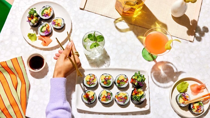 Overhead shot of two plates of veggie sushi rolls with chopsticks, soy sauce, and cocktails on a white marble table.