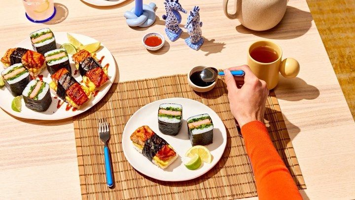 Overhead shot of sushi dishes, condiments, and drinks on a wooden table, with a hand adding soy sauce to one of the sushi pieces.