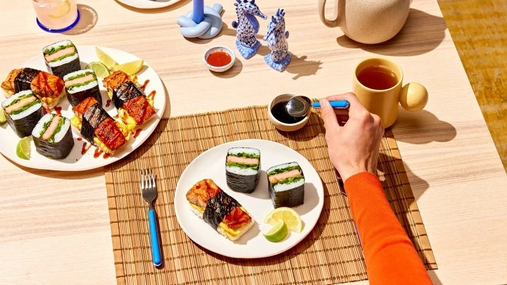 Overhead shot of sushi dishes, condiments, and drinks on a wooden table, with a hand adding soy sauce to one of the sushi pieces.