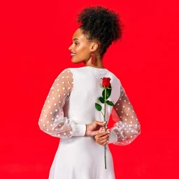 Woman in white dress with sheer polka dot sleeves holding a red rose behind her back against a red background