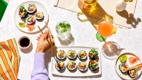 Overhead food photography of sushi rolls, soy sauce, cocktail, and chopsticks held by a person in a lavender sweater.