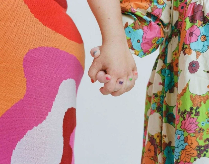 Two women holding hands. One woman wears a dress with pink, orange, and white wavy patterns. The other wears a white floral-patterned dress.