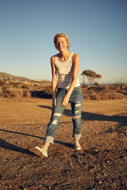 A blonde woman stands in ripped jeans and a tank top, smiling in a sunny outdoor environment with scrubby vegetation in the background.