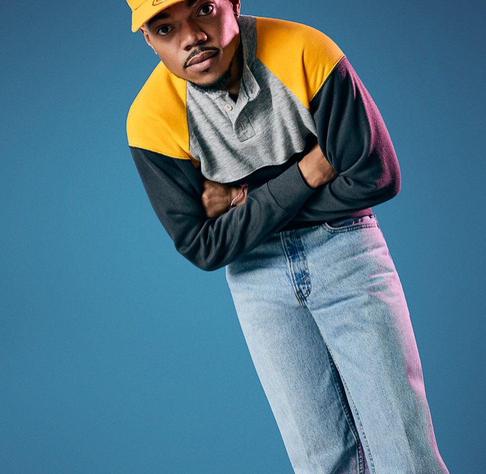 Portrait of a man with arms crossed wearing a yellow hat and multicolored long sleeve shirt against a blue studio background.