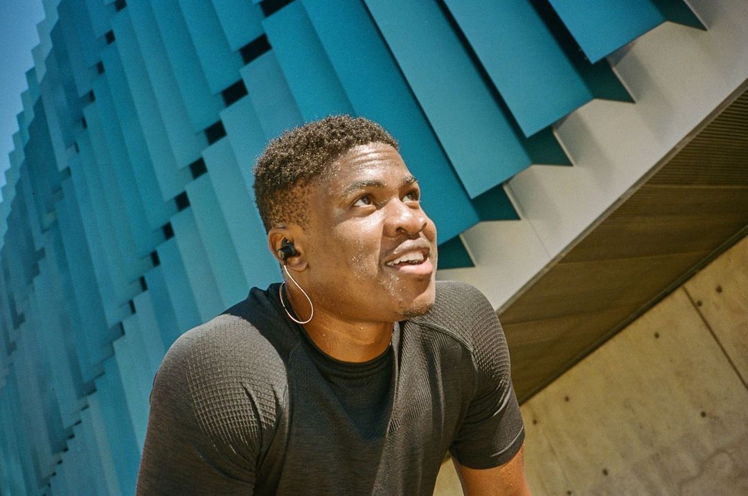 Low-angle shot of a sweaty Black adult male with athletic wear and earbuds outside a modern building during the day.