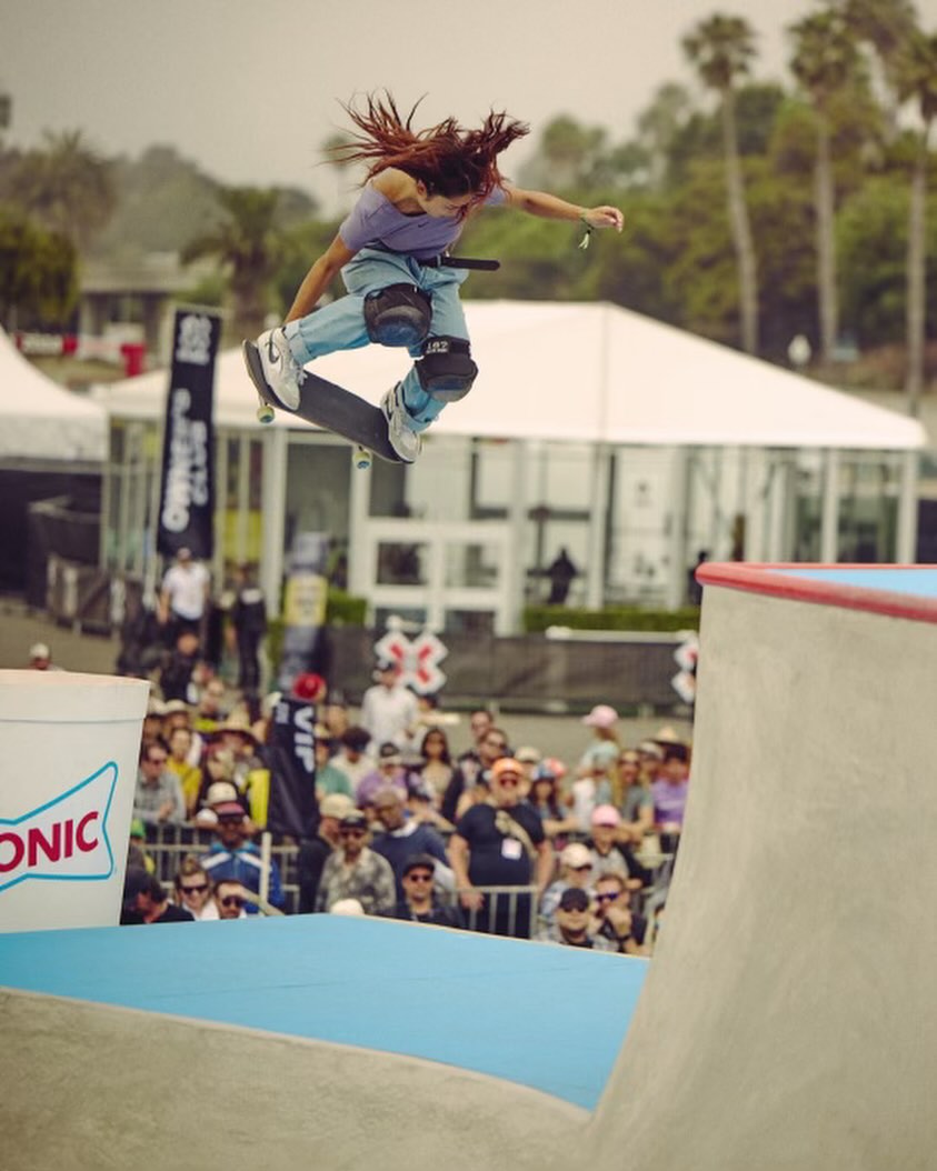 Female skateboarder mid-air performing a trick on a skateboard ramp. Crowd watches from the background at an outdoor event.