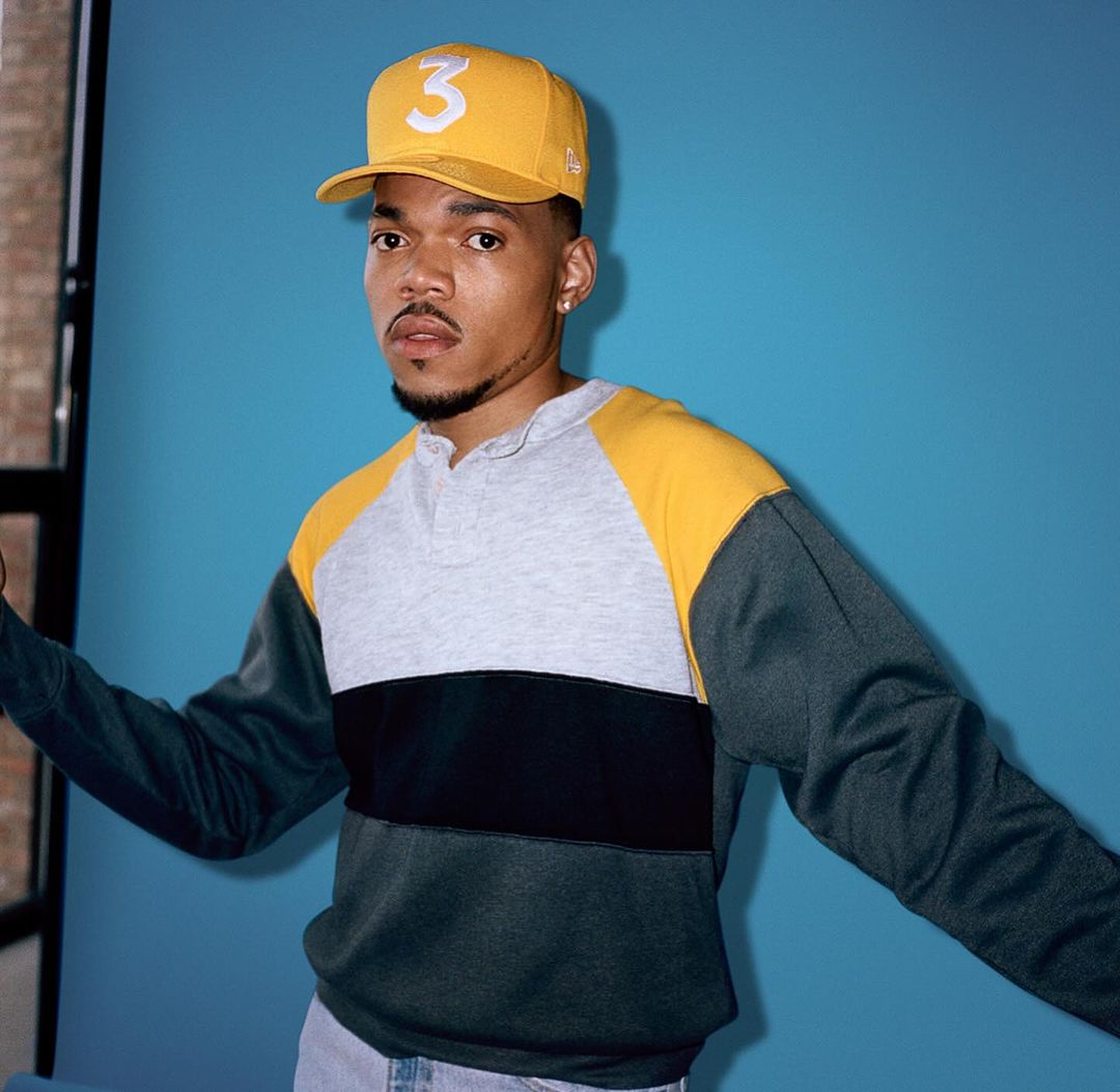 Portrait of a young adult male wearing a yellow baseball cap with a '3' logo, a color-blocked sweatshirt, and jeans against a blue backdrop.