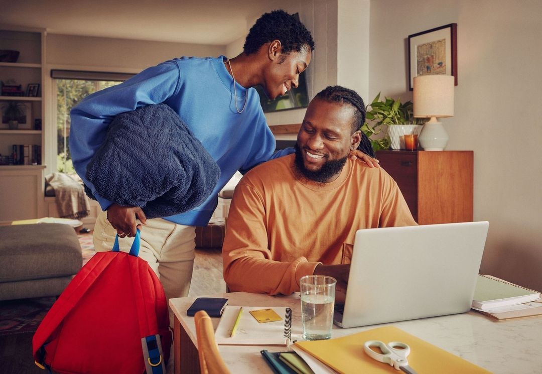 A happy African-American couple at home. The woman carries a bag, while the man works on his laptop at a table with papers and office supplies.