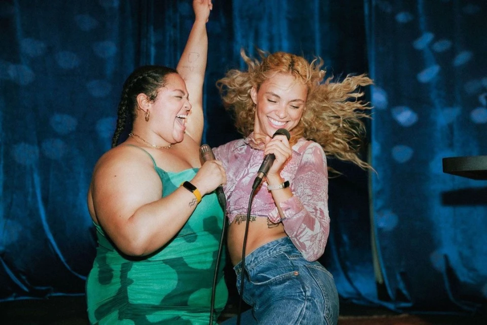 Two adult females sing into microphones on stage in front of a blue curtain with spot lighting, wearing fashionable outfits, and yellow wristbands.