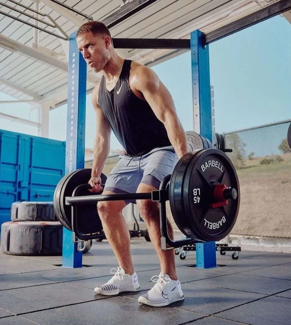 Muscular man lifting barbell with weights in gym, wearing tank top, shorts, and sneakers. Fitness training and workout.