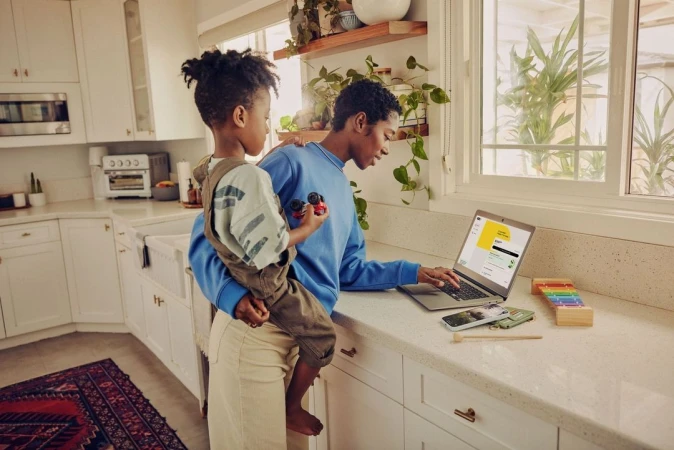 Woman and child in a kitchen, woman working on a laptop, child holding a toy car, domestic scene with natural light and indoor plants.