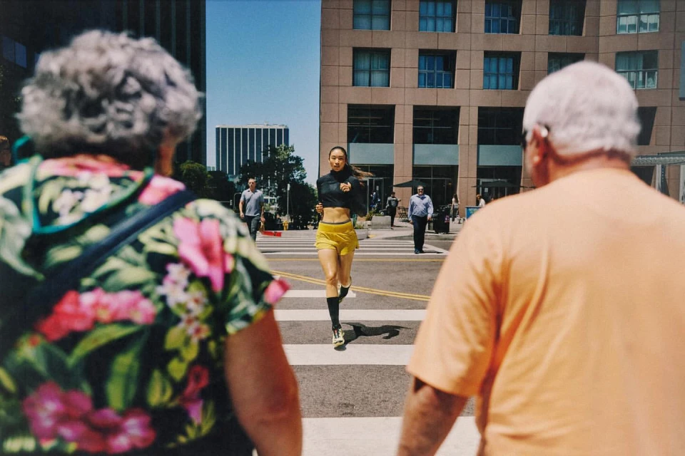 Woman in athletic wear runs across a crosswalk in a city setting with buildings, pedestrians, and bright daylight.