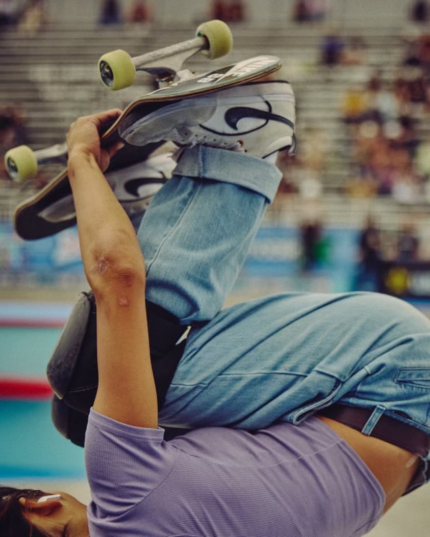 Action shot of a skateboarder performing a trick indoors, showcasing a close-up of the skateboard, shoes, and the athlete's arm and elbow.