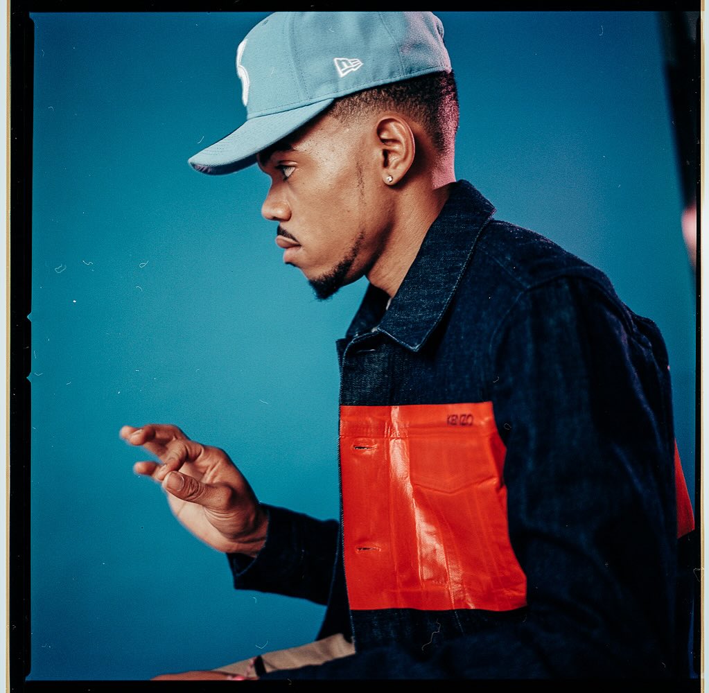Portrait of an African American man in a baseball cap and denim jacket with red accents, against a light blue backdrop in studio.