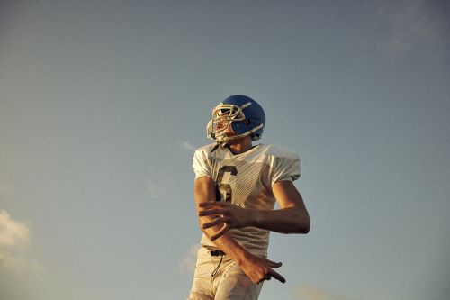 Adult male in sports uniform and helmet outdoors against a light-blue sky during daytime, in possible action pose, focused on the game.