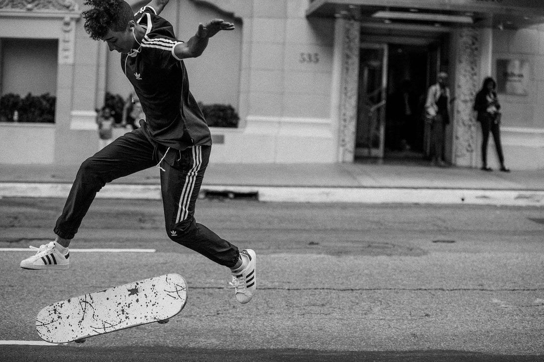 Teenager jumps over a skateboard on a city street in black and white, wearing Adidas apparel, with an urban building in the background.