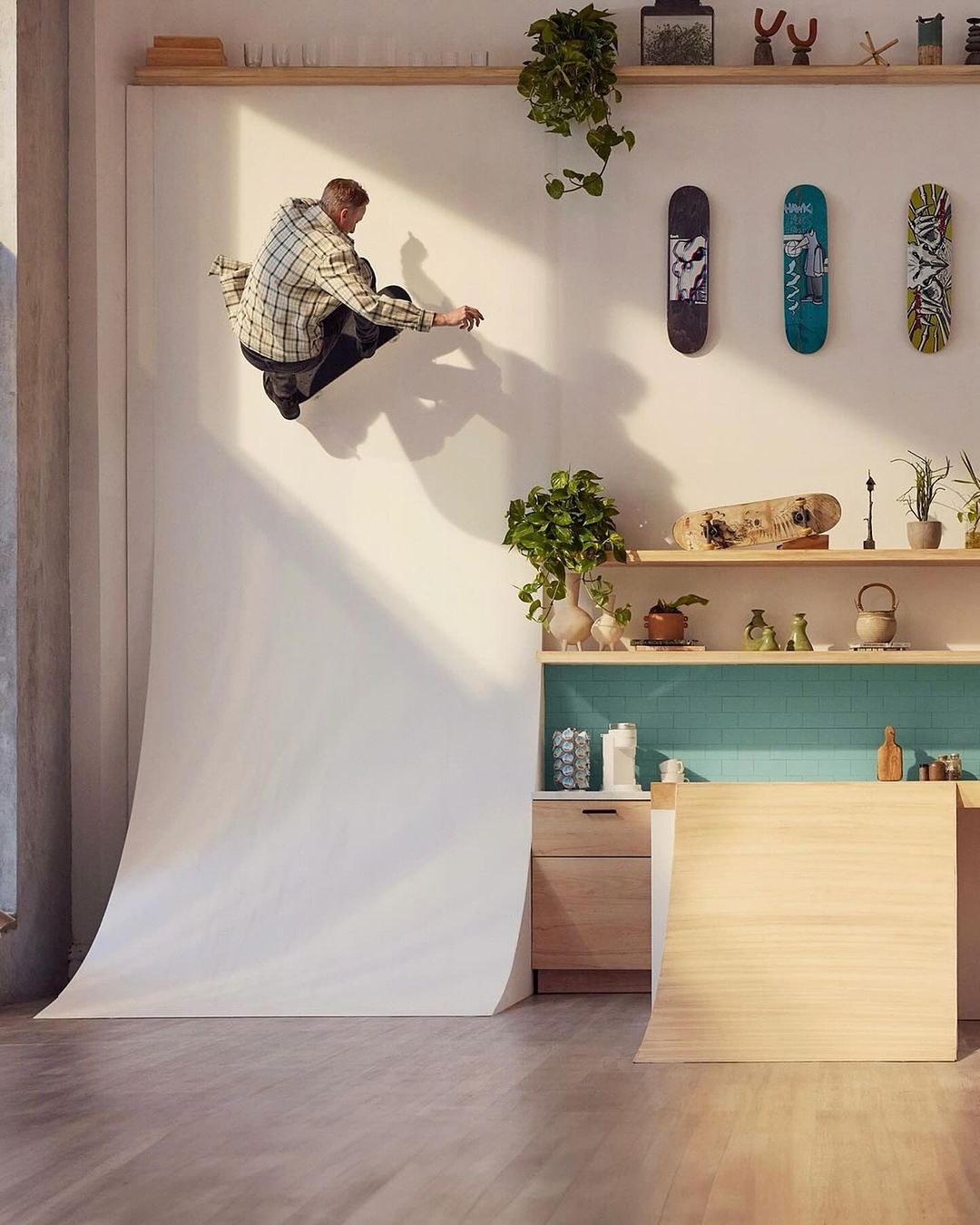 An adult male skateboarder crouches mid-air on a white ramp in an indoor room with plants and skateboards displayed on a shelf.