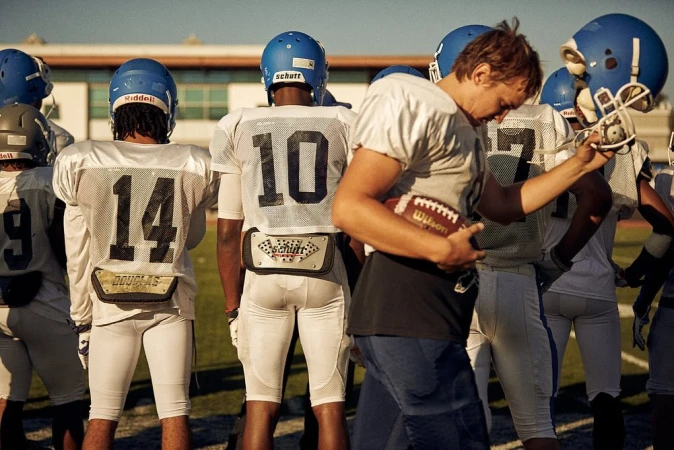 American football players in uniform stand on the field in the daytime with a person holding the ball in the foreground.