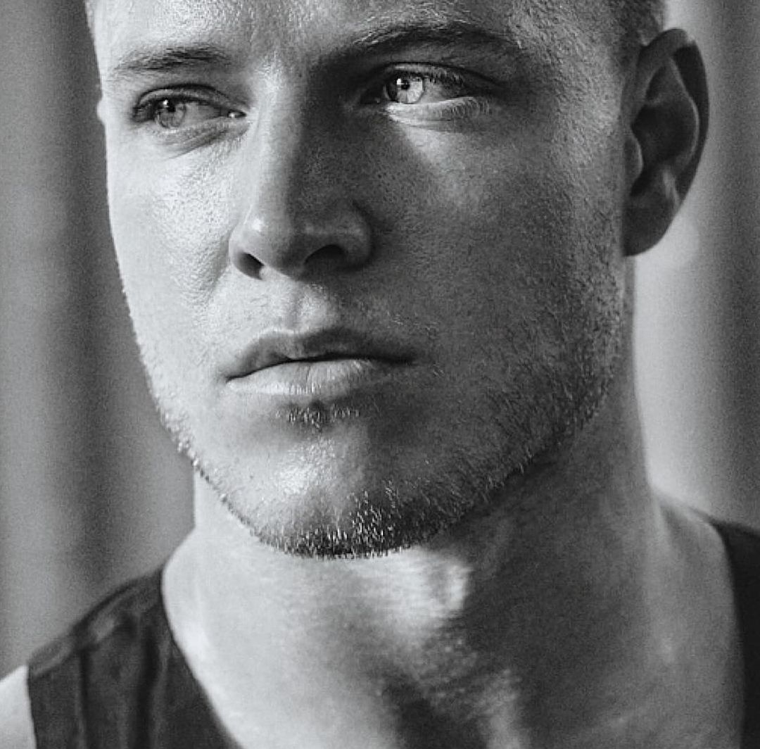 Close-up monochrome portrait of an adult male with short hair and a slight beard, looking off-camera with a neutral expression.