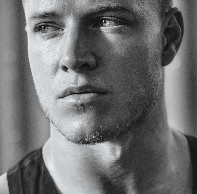 Close-up monochrome portrait of an adult male with short hair and a slight beard, looking off-camera with a neutral expression.