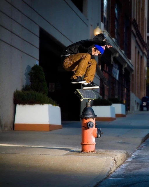 A skateboarder balances on a board placed on top of an orange fire hydrant on a city street, captured in a medium outdoor shot.