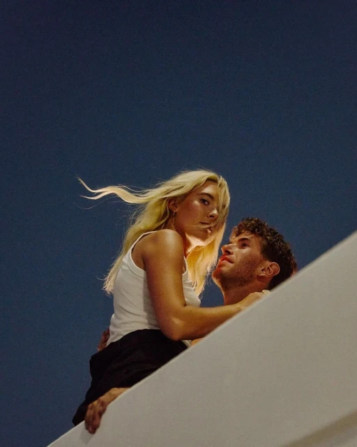 Close up of a blonde woman embracing a man in dark clothing against a dark sky, creating a romantic and intimate atmosphere.