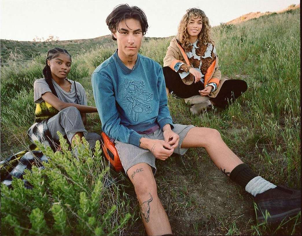 Three people are sitting on a grassy hillside. Two are light skinned, one is dark skinned. All are wearing casual clothing on a sunny day.