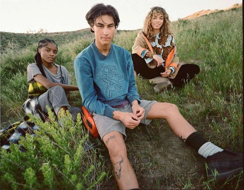 Three people are sitting on a grassy hillside. Two are light skinned, one is dark skinned. All are wearing casual clothing on a sunny day.