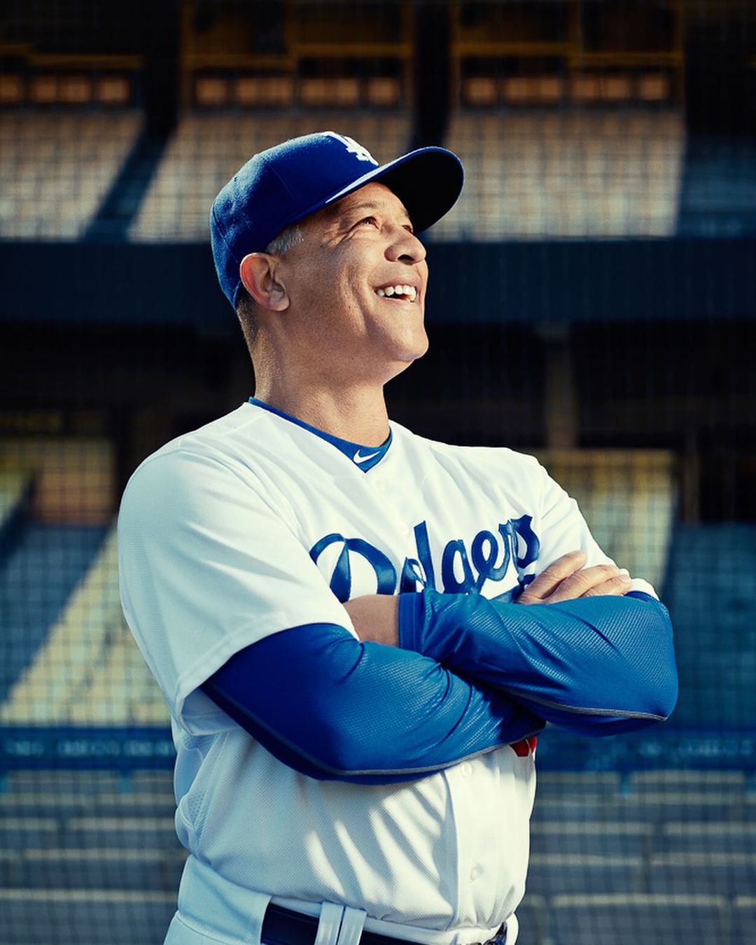 An adult man smiles wearing a baseball uniform and hat with the Dodgers logo on a baseball field with stadium seating in the background.
