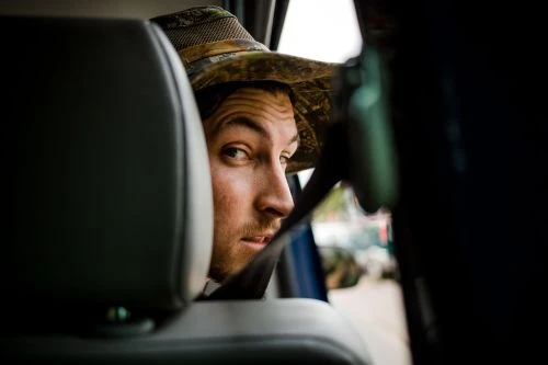 Man looks back from car seat, wearing camo hat and seatbelt, creating an eye-level portrait in natural light.