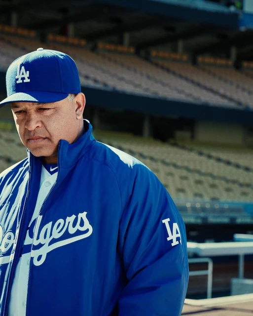 Adult male wearing a blue LA Dodgers baseball cap and jacket stands in a baseball stadium with blue seating as the background.