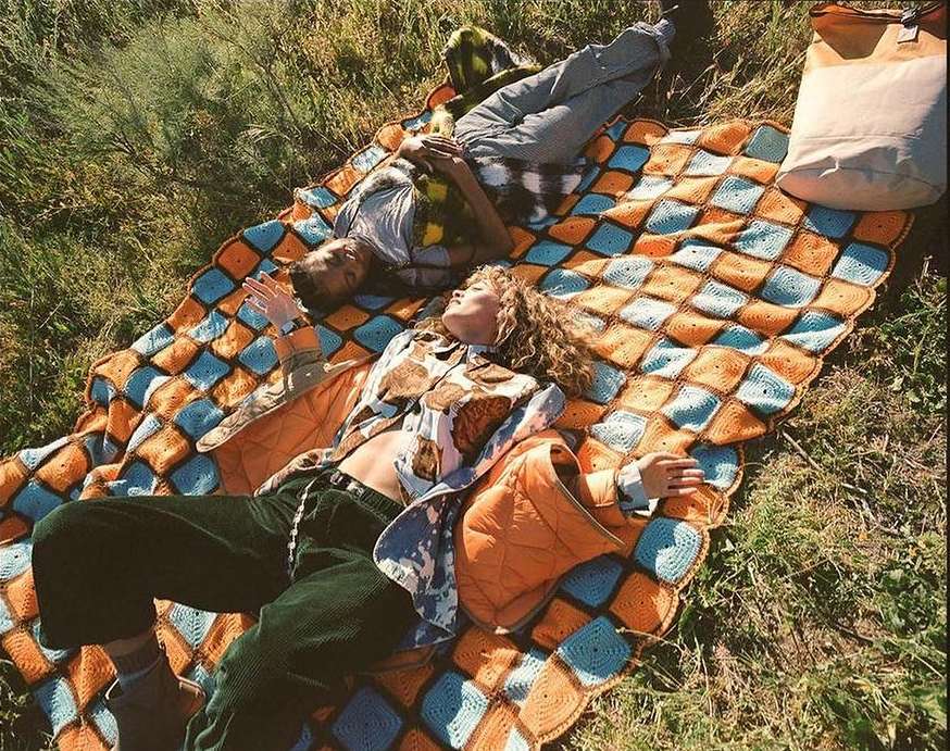 Two people relax on a patterned blanket outdoors in a grassy area, with one individual on their back looking up at the sky.
