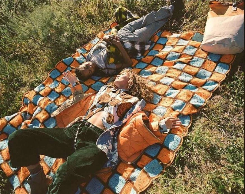 Two people relax on a patterned blanket outdoors in a grassy area, with one individual on their back looking up at the sky.