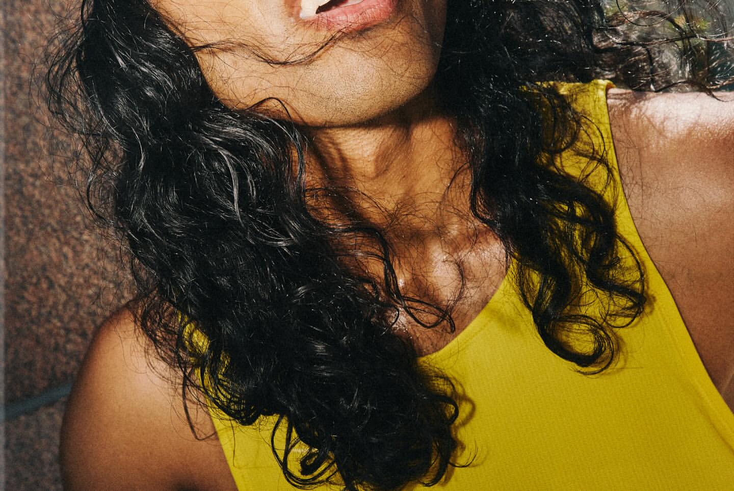 Close-up shot of an adult with long curly dark hair and wearing a bright yellow tank top, with a textured background visible.