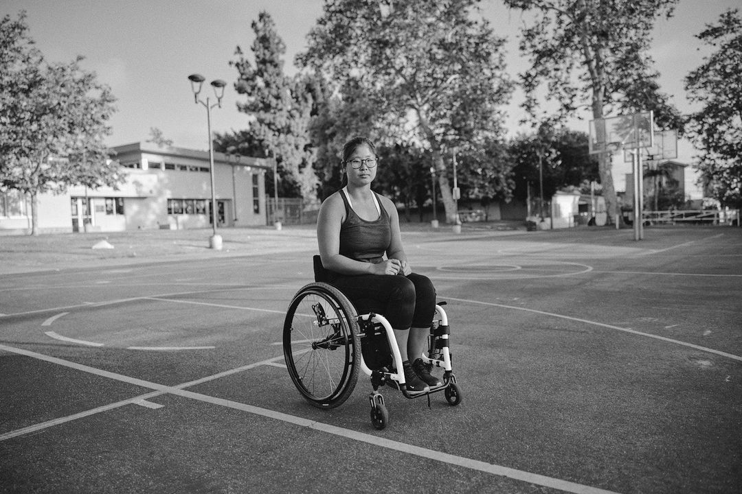 Portrait of a woman sitting in a wheelchair in a black and white outdoor setting on a basketball court with trees and building.