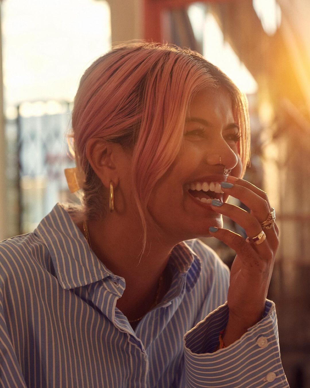 Smiling woman with pink hair wearing a striped shirt and gold jewelry. Indoor shot with natural light illuminating the subject's face.