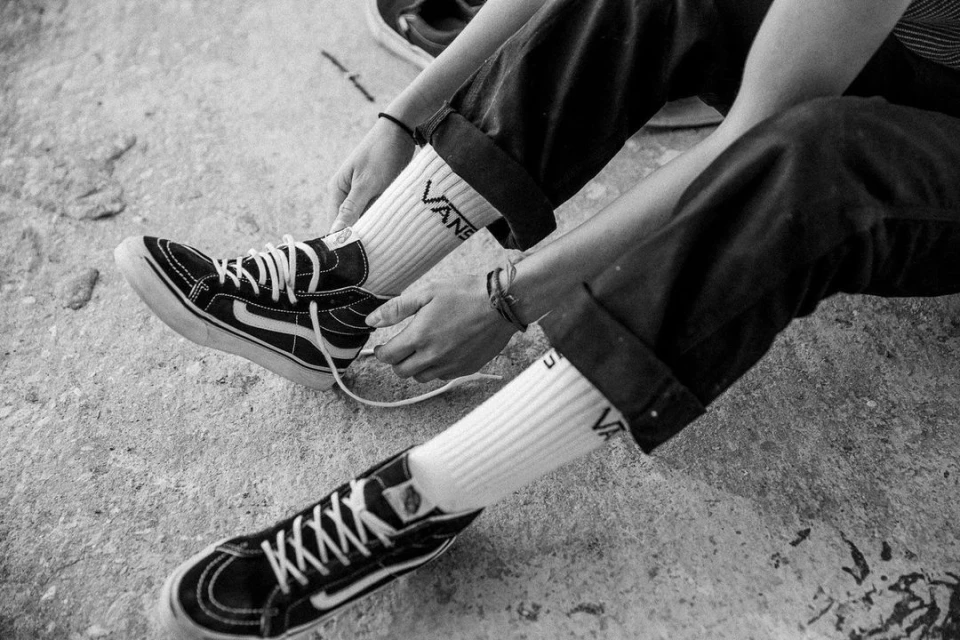 A person is sitting and tying the laces of a pair of Vans sneakers while wearing socks with the brand's logo; the image is in black and white.