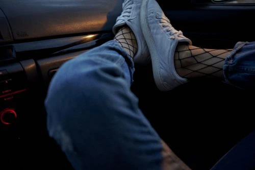 Legs in denim jeans and white sneakers with fishnet socks on dashboard inside car interior at nighttime.