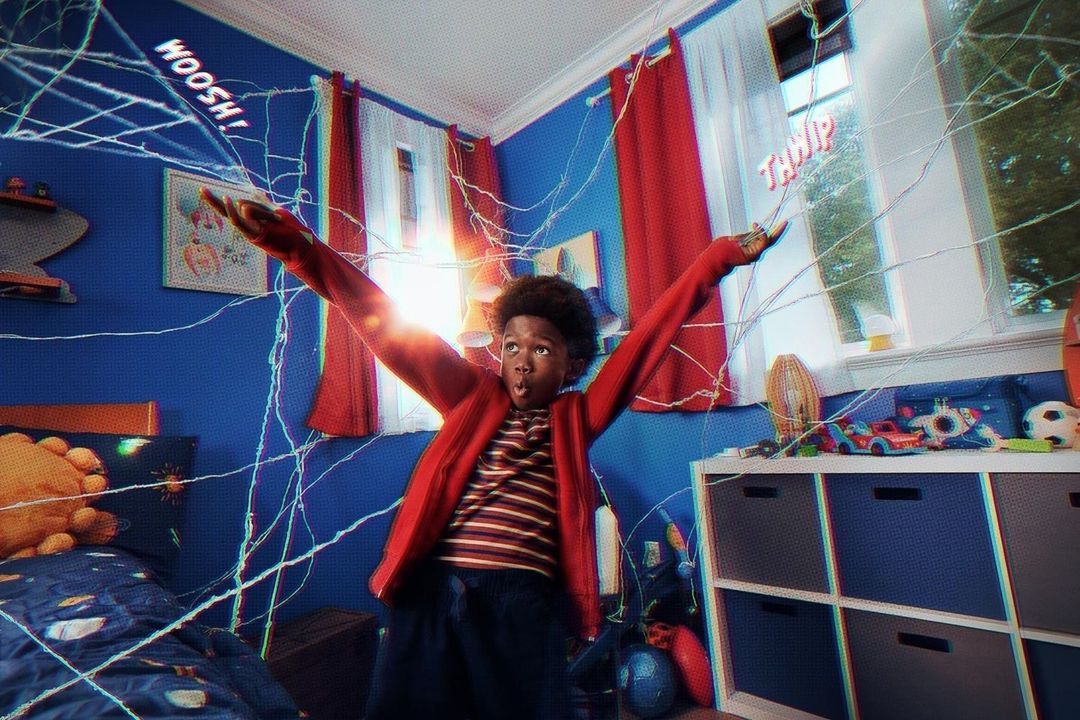 Boy with arms raised in a web-covered bedroom. He wears a red jacket and looks surprised, surrounded by an indoor, playful setting.