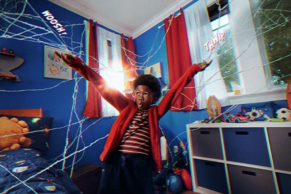 Boy with arms raised in a web-covered bedroom. He wears a red jacket and looks surprised, surrounded by an indoor, playful setting.